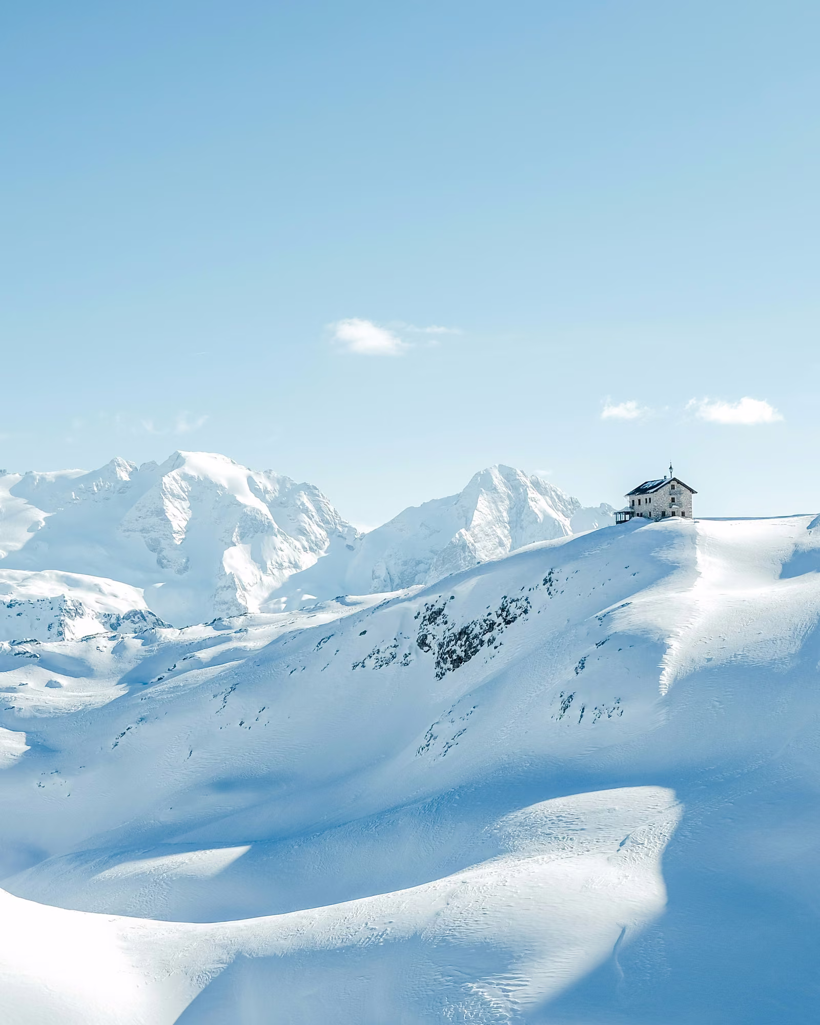 A mountain refuge stands above broad snow-covered slopes and high Alpine peaks.